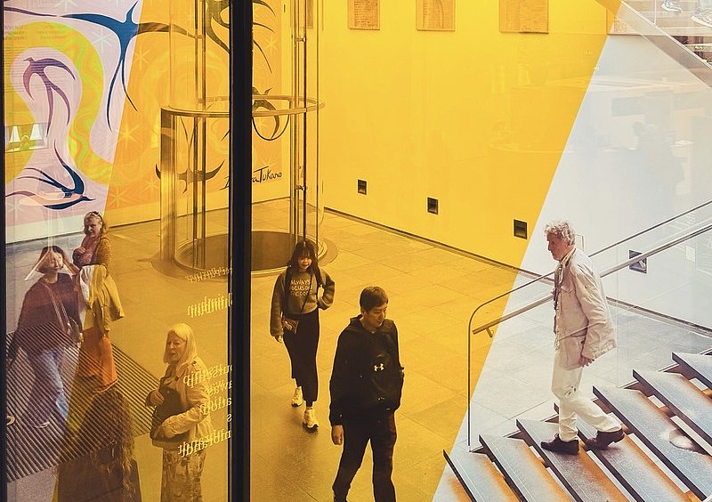 Besucherinnen und Besucher im Foyer des Mauritshuis, mit gelber Glasfläche, gläsernem Aufzug und Treppe.