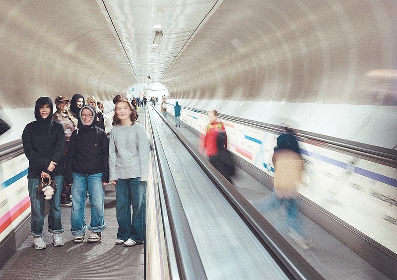Schülerinnen auf einem Fahrsteig in einem hellen Tunnel; Menschen auf dem benachbarten Fahrsteig erscheinen bewegungsunscharf.