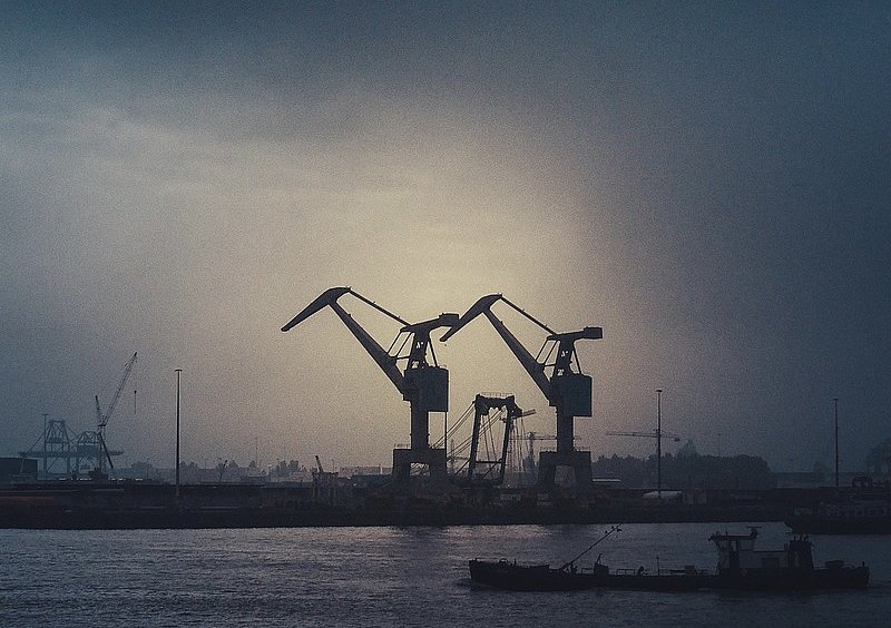  Zwei Hafenkräne als dunkle Silhouetten vor der diesigen Sonne, davor ein Schiff auf dem Wasser.