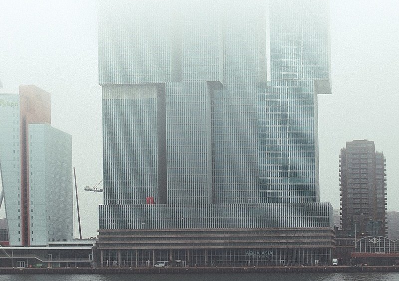  Die versetzten gläsernen Hochhaustürme von De Rotterdam am Wasser, deren obere Stockwerke im Nebel verschwinden.