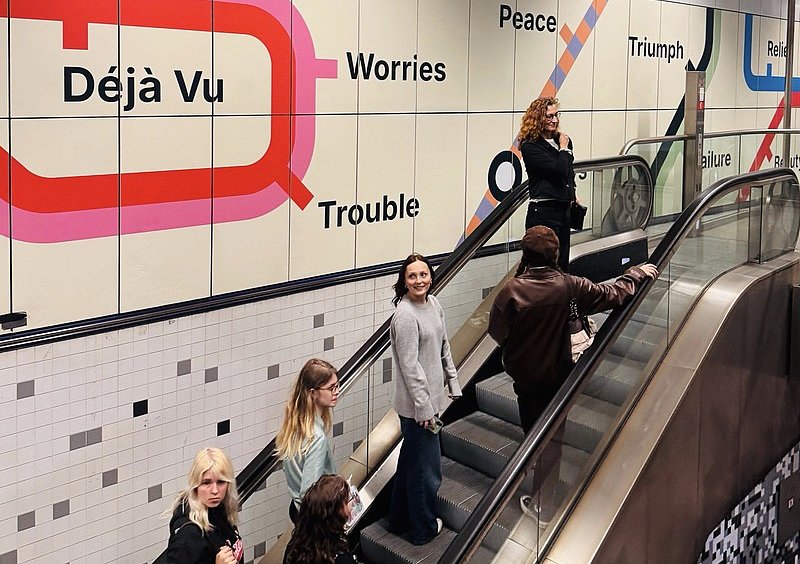 Schülerinnen auf einer Rolltreppe neben einer Wandgestaltung mit bunten Linien und englischen Begriffen wie „Déjà Vu“ und „Worries“.